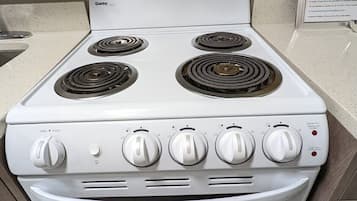 Una estufa eléctrica Danby blanca con cuatro hornillas y un horno, ubicada en una cocina con encimera color beige y un letrero en la pared.