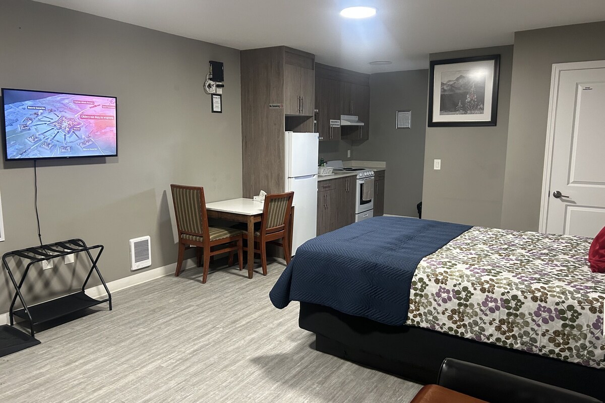 A hotel room with a bed, a desk, a chair, a kitchenette, and a television displaying a map.