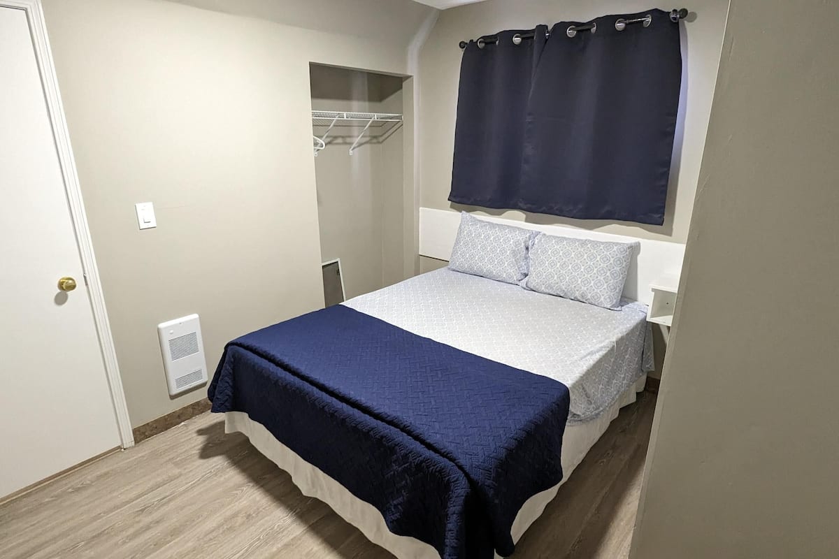 Um quarto com cama, guarda-roupa, janela com cortinas e um respiradouro.