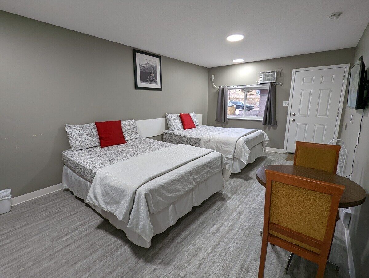 A room with two beds, a chair, a table, a door, and a picture on the wall.