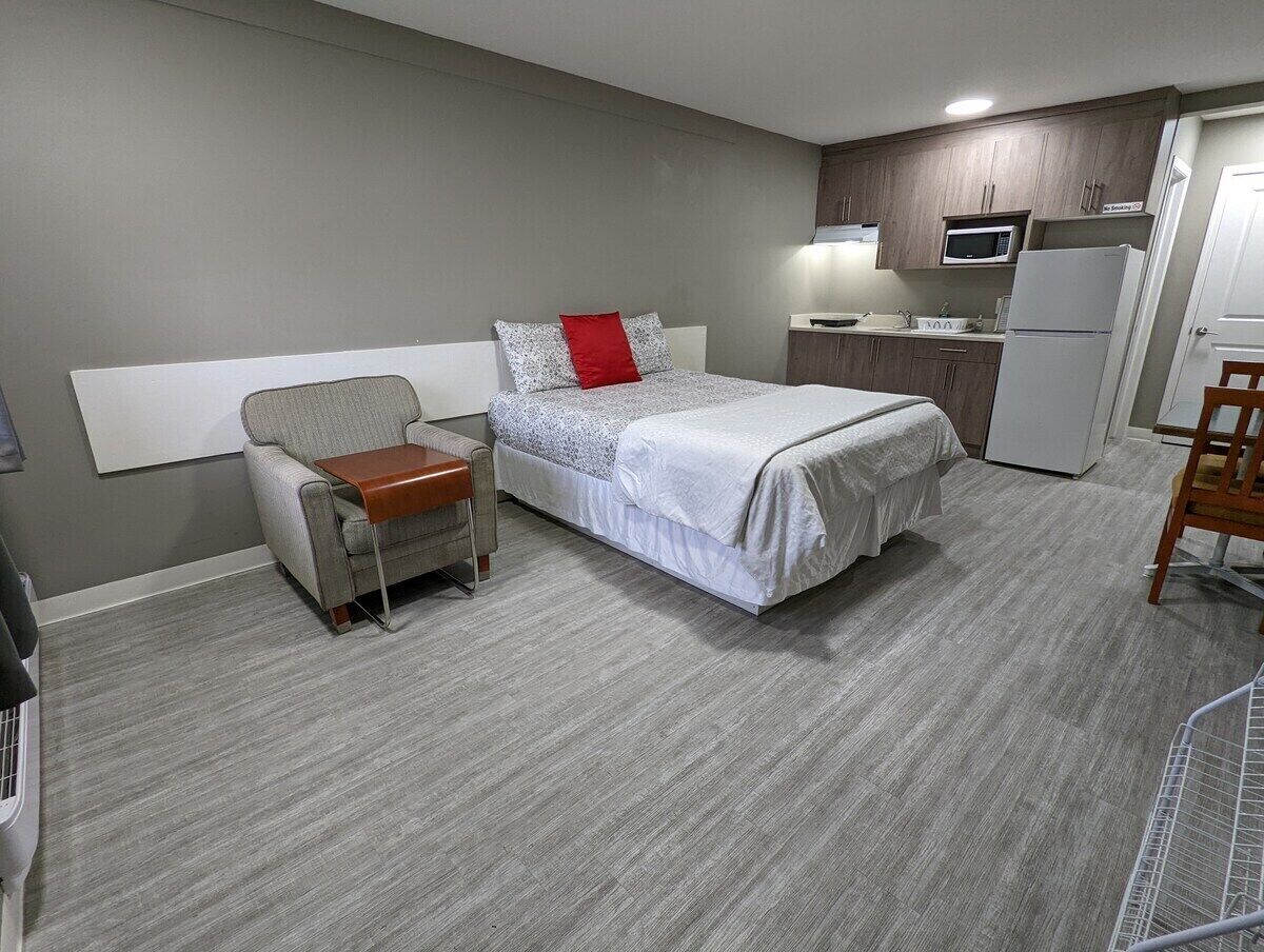 A hotel room with a bed, a chair, a small table, a microwave, a refrigerator, and a kitchenette.