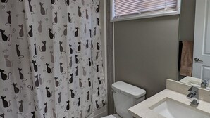 A bathroom with a toilet, a sink, a shower curtain with cat patterns, a window with blinds, and a towel rack. - Riverside Motel & Suites (Medicine Hat)