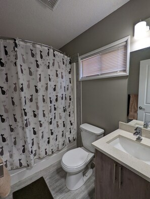 A bathroom with a toilet, a sink, a shower curtain with cat patterns, a window with blinds, and a towel rack. - Riverside Motel & Suites (Medicine Hat)