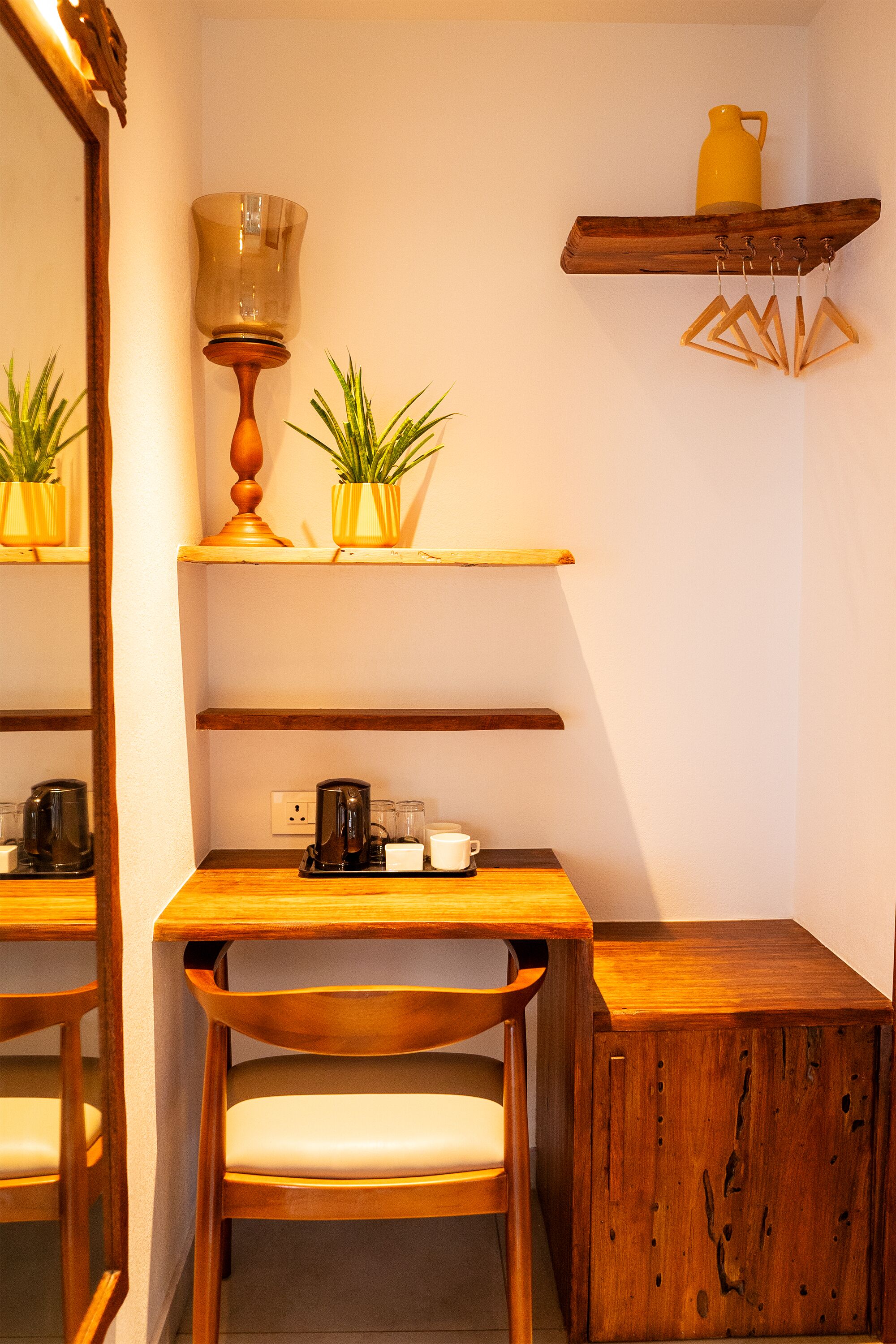 Un escritorio de madera con una silla, un espejo y una estantería con plantas y una jarra amarilla.