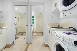 A modern bathroom with a glass-enclosed shower, a washing machine, and a toilet visible through the glass door. - Beach Club 5113/4 | 2 Bedroom Pool Access Apartment (Palm Cove)
