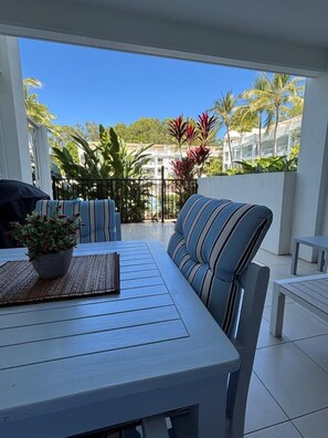A patio with a striped sofa, a table with a plant, and a view of a residential area with palm trees and houses. - Beach Club 5113/4 | 2 Bedroom Pool Access Apartment (Palm Cove)