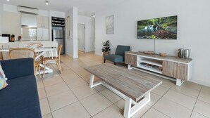 A modern living room with a sofa, a coffee table, a TV, and a dining area with chairs and a table. - 2 Bedroom Ground Floor Pool Access Apartment 5113/4 (Palm Cove)