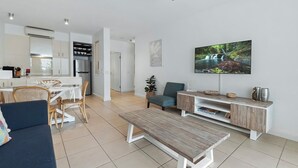 A modern living room with a sofa, a coffee table, a TV, and a dining area with chairs and a table. - 1 Bedroom Ground Floor Pool Access Apartment 5114 (Palm Cove)