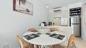 A modern kitchen with white cabinets, a stainless steel refrigerator, and a dining area with a white table and wicker chairs. - 1 Bedroom Ground Floor Pool Access Apartment 5114 (Palm Cove)
