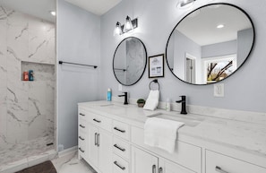 A modern bathroom with a white marble vanity, two round mirrors, a towel rack, and a shower area. - Inviting 4BR 2 5BA Downtown Home With Ample Space (San Antonio)