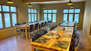 Une longue table à manger entourée de chaises, de grandes fenêtres et des luminaires suspendus.