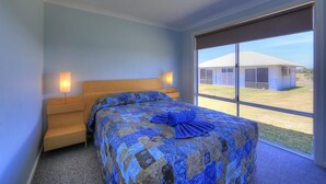 A bedroom with a large bed, a wooden headboard, two bedside lamps, and a view of a building through a large window. - Tasman Holiday Parks - Ledge Point (Ledge Point)