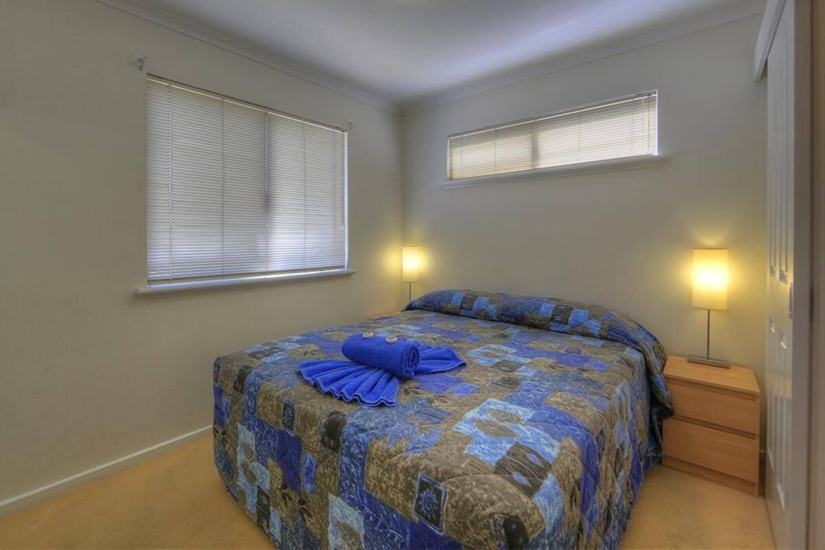 A bedroom with a bed, two bedside lamps, and two windows with blinds.