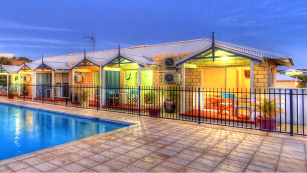A swimming pool with lounge chairs and a row of brightly lit cottages. - Tasman Holiday Parks - Ledge Point (Ledge Point)