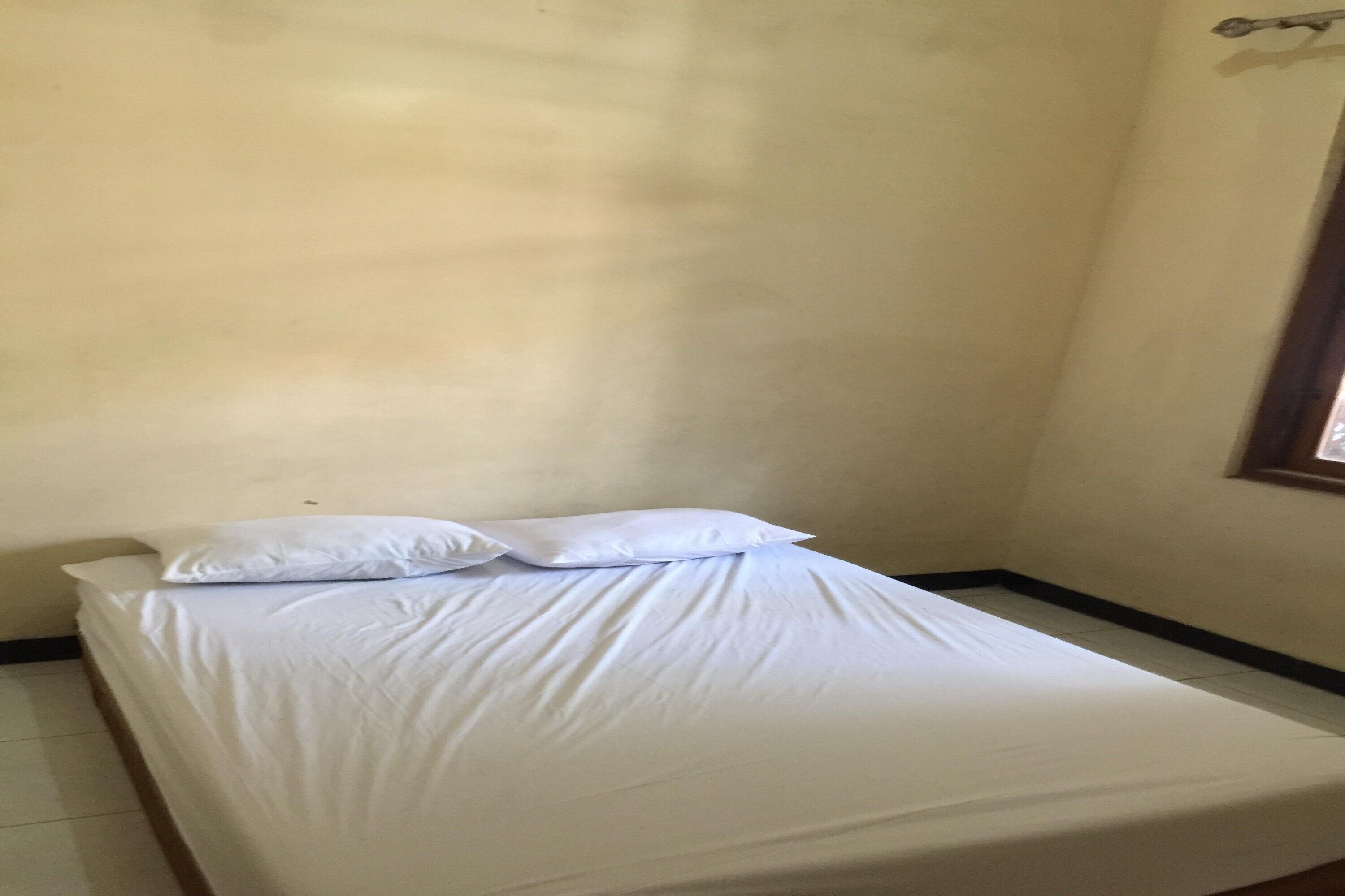 Una cama individual con ropa de cama blanca en una habitación con paredes lisas y una ventana a la derecha.