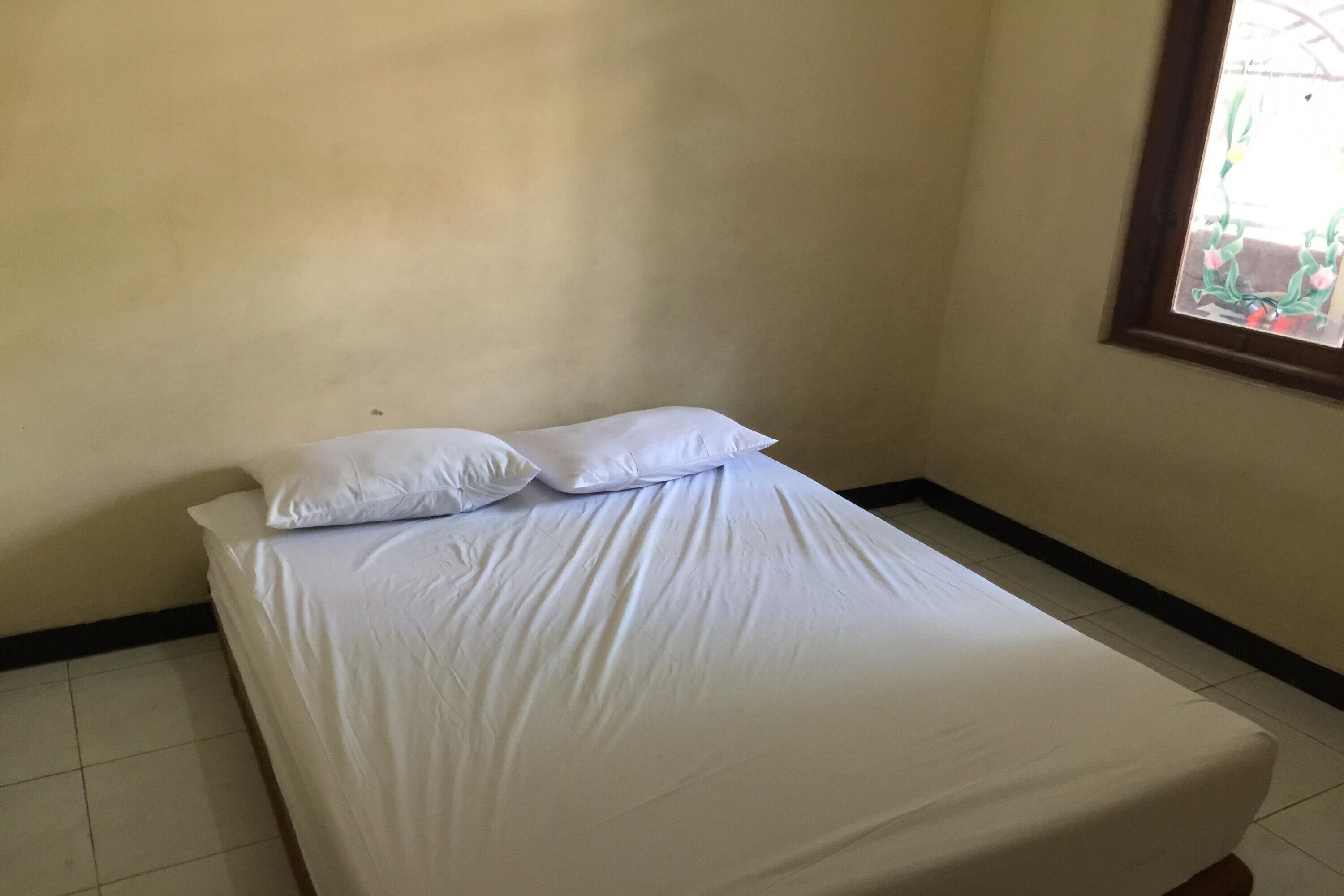 A single bed with white bedding in a room with plain walls and a window with a view of greenery.