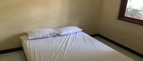 A single bed with white bedding in a room with plain walls and a window with a view of greenery.