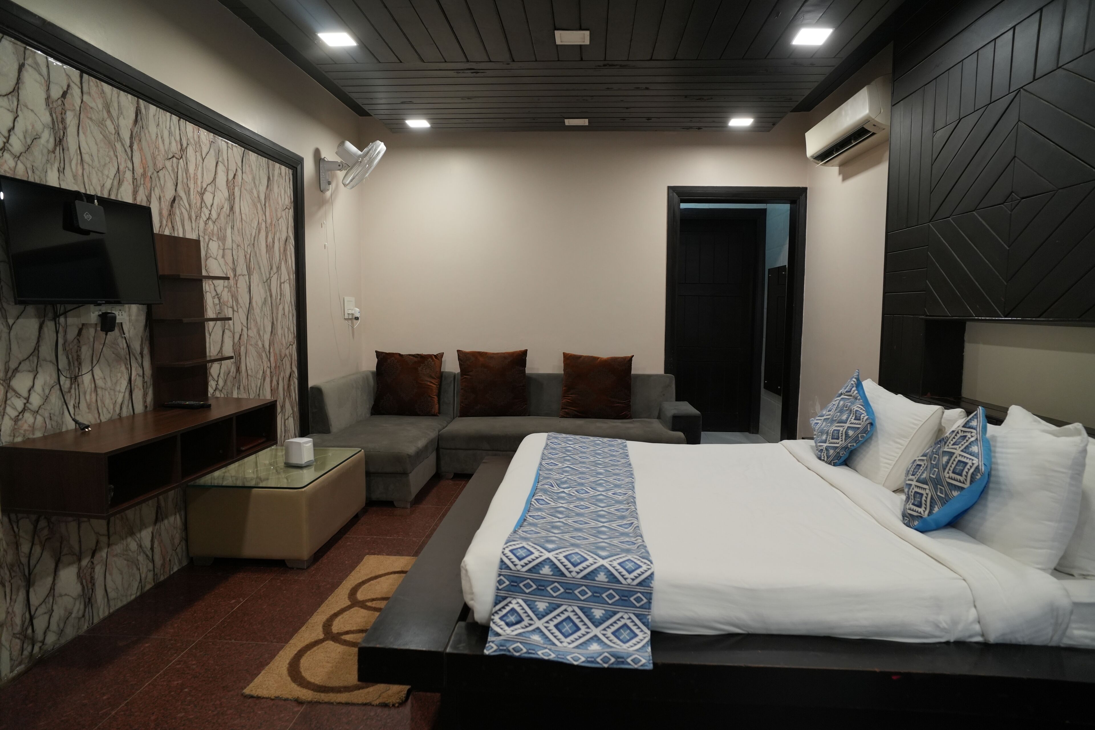 A modern bedroom with a large bed, a sofa, a TV mounted on a marble wall, and a small table with a lamp.