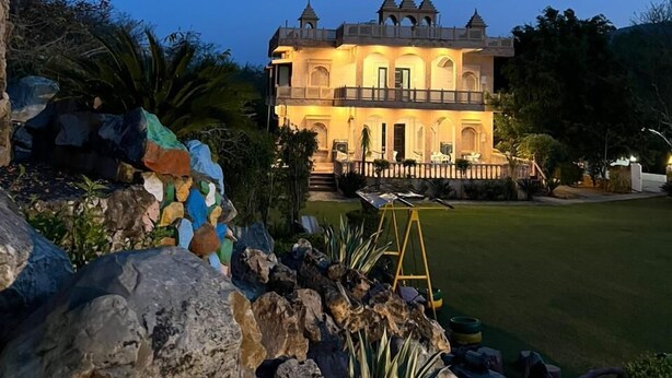 Un edificio grande de varios pisos, con un balcón y ventanas arqueadas, rodeado por un jardín ajardinado con rocas y plantas.