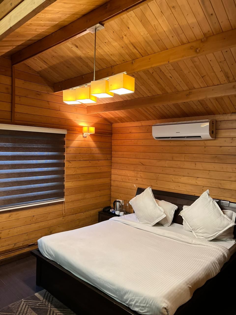 A wooden-paneled room with a bed, a window with blinds, a wall-mounted air conditioner, and a hanging light fixture.