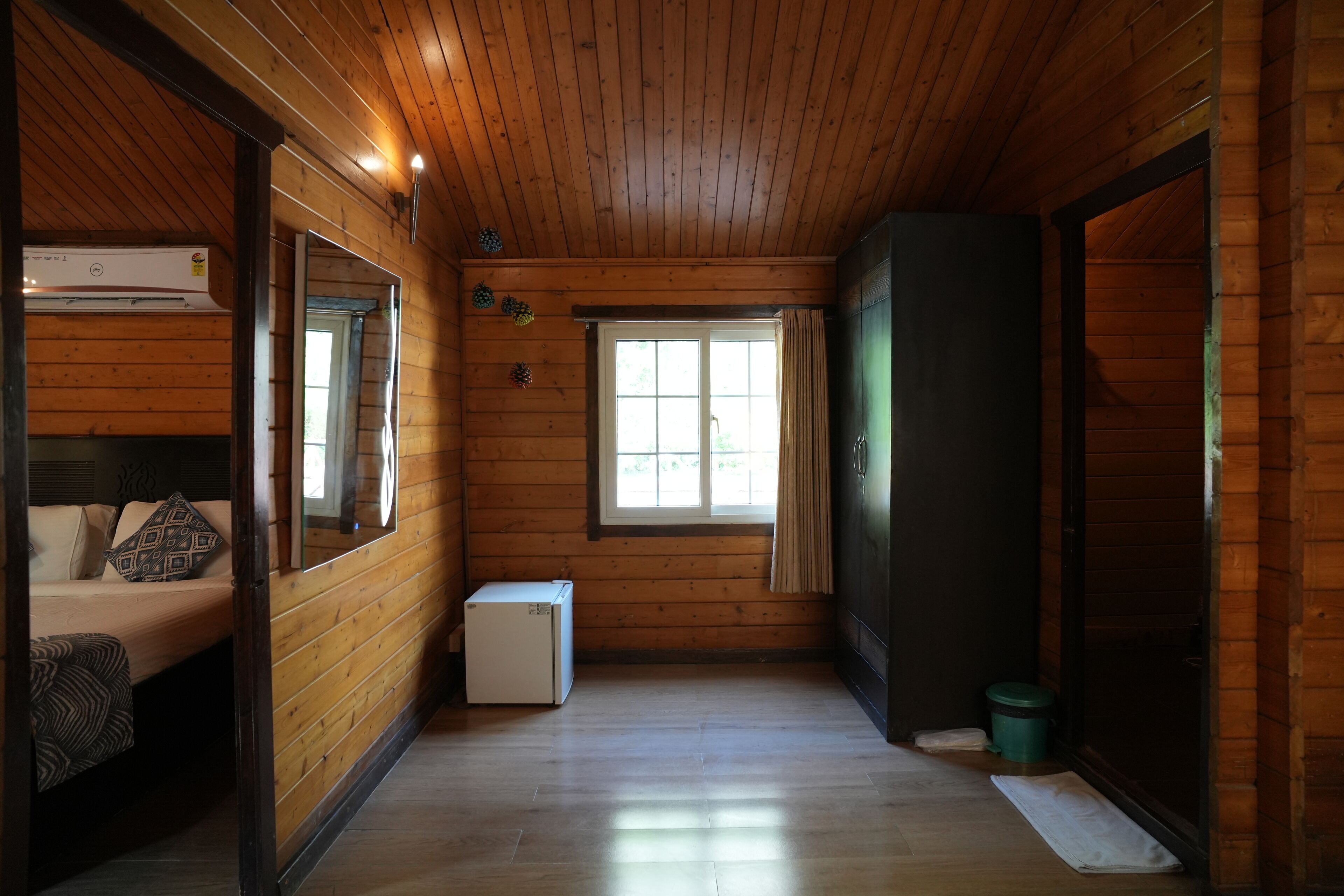 A cabin interior with wooden walls, a bed with patterned pillows, a small white refrigerator, a window with curtains, and a door leading to another room.