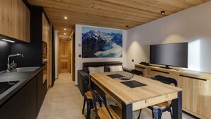 A modern kitchen with a wooden ceiling, a dining area with a wooden table and chairs, a black sofa, and a large TV on the right. - Vouvy - Moderne 4pax | Remontée Flégère 5min | Praz | Arve (Chamonix-Mont-Blanc)