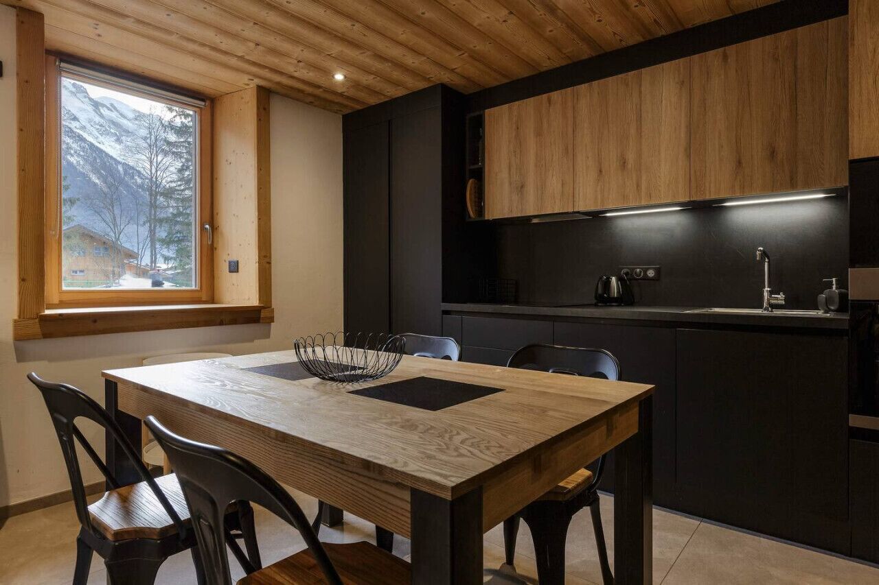 Une cuisine moderne avec une table et des chaises en bois, une fenêtre donnant sur les montagnes et des meubles de rangement aux couleurs sombres.