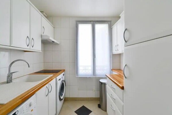 A compact kitchen with white cabinets, a wooden countertop, a washing machine, and a window with blinds. - The Cosy Cocon -Close to Eiffel- by Mon Petit Faré (Paris)