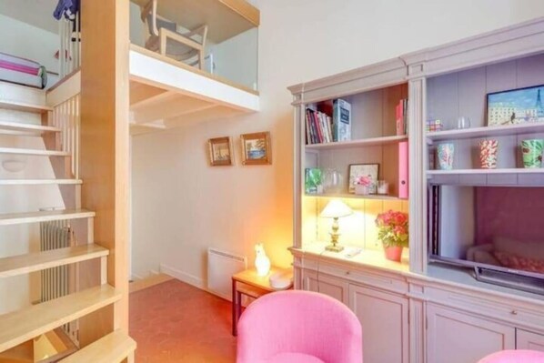 A room with a loft bed, a bookshelf, a small table with a lamp, and a pink chair.