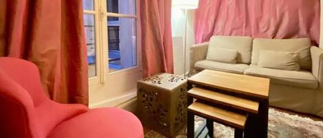 Una habitación con una silla roja, un sofá color beige, una pequeña mesa de madera y una alfombra estampada.