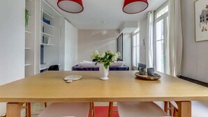 Ein Essbereich mit Holztisch, zwei roten Pendelleuchten, einer Blumenvase und Blick auf ein Schlafzimmer mit Bett und Vorhängen.