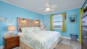 A bedroom with a bed, nightstands, a ceiling fan, a window with curtains, a mirror, and a trash can. - 5201 - Flip Flop Time - Cozy Oceanfront Condo | OBX Getaway (Kill Devil Hills)