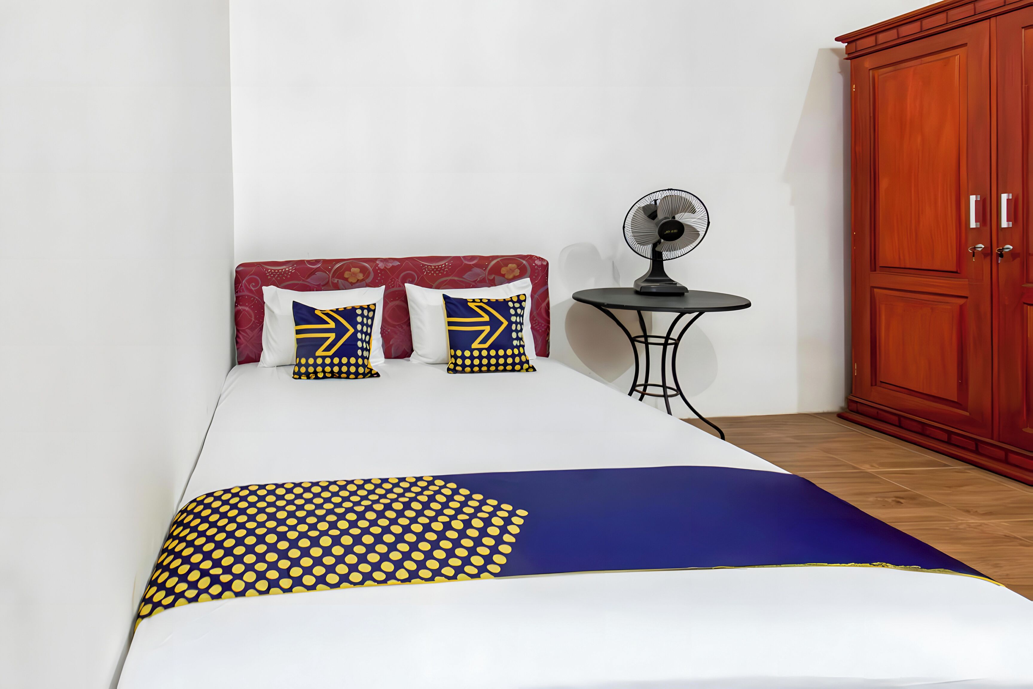 Ein Bett mit gemustertem Überwurf, zwei Kissen mit geometrischem Muster, ein kleiner Tisch mit Ventilator und ein Holzschrank.