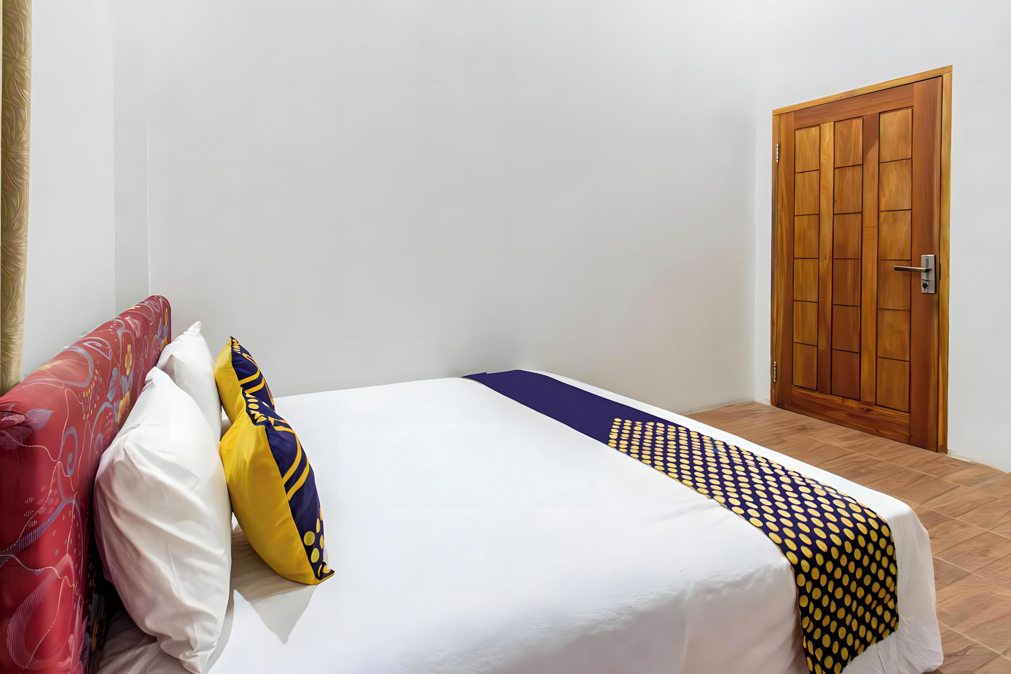 Una cama bien hecha con una colcha blanca y estampada, un cojín rojo y amarillo, y una puerta de madera al fondo.