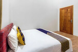 Una cama bien hecha con una colcha blanca y estampada, un cojín rojo y amarillo, y una puerta de madera al fondo.