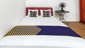 A bed with a patterned bedspread, two pillows with geometric designs, a bedside table with a fan, and a wooden wardrobe. - Hotel O Kebun Manis Homestay Near Lapangan Bola Volly Batu Kumbung (Janapria)