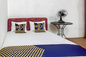 Una cama con una colcha estampada, dos almohadas con diseños geométricos, una mesita con un ventilador y un cabecero rojo.