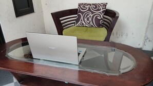 A laptop on a glass-top wooden table, a chair with a patterned cushion, and a partially visible wall.