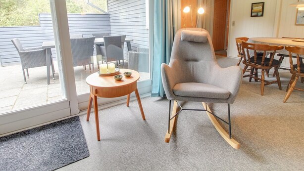 A room with a large sliding glass door leading to an outdoor patio area. Inside, there is a grey rocking chair, a wooden dining table with chairs, and a small round table with candles and bowls. - "Sonia" - 3km from the sea by Interhome (Nørre Nebel)