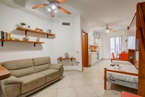 A modern living room with a sofa, wooden shelves, a ceiling fan, and a view into the kitchen area. - Verbena with parking 500m from the sea (Alassio)