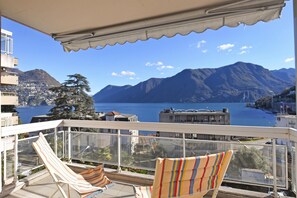 A balcony with two striped lounge chairs, a glass railing, and a view of mountains and a body of water. - Sweet Lake View Lugano (Lugano)