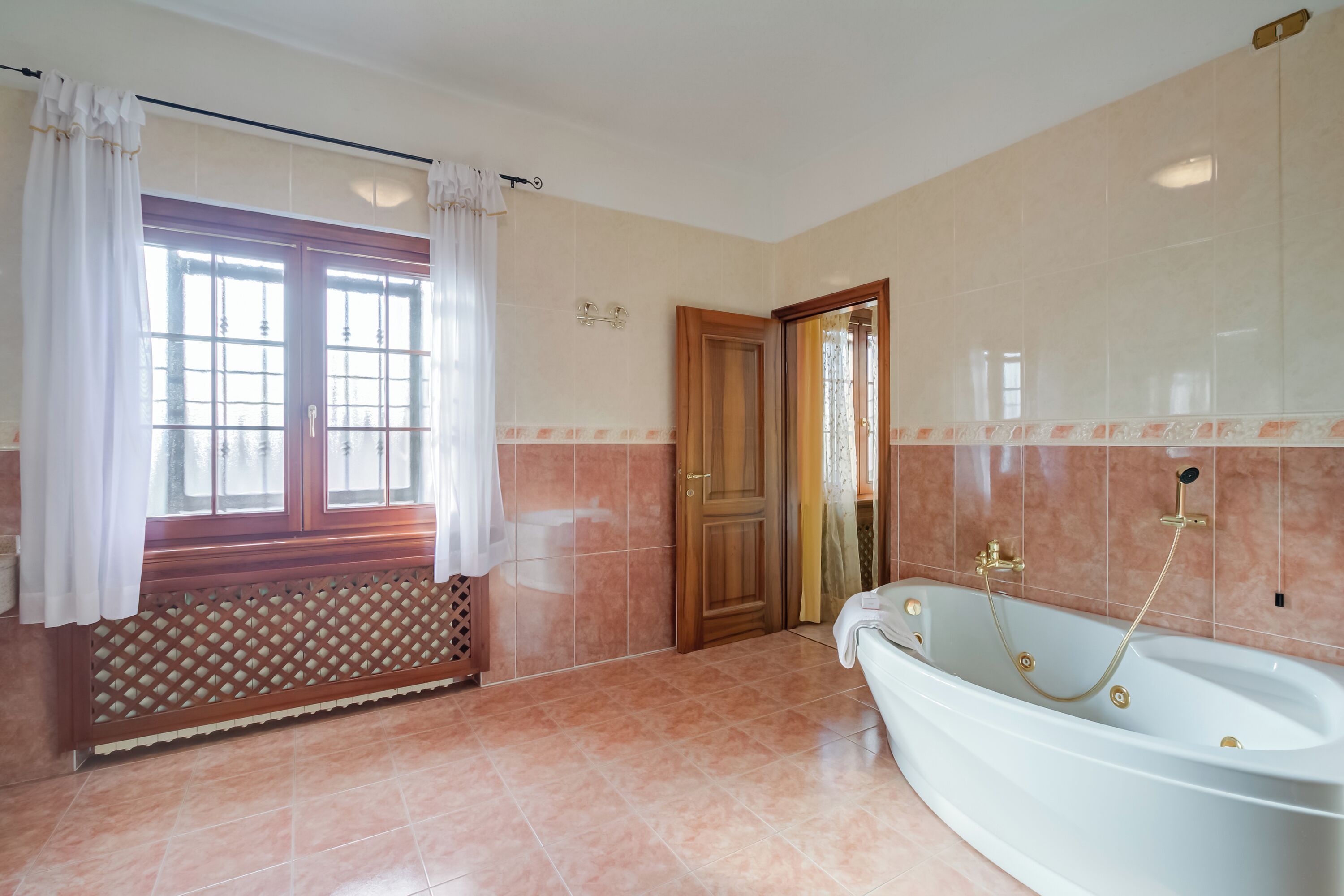 Uma casa de banho com uma banheira, uma janela com cortinas, um radiador e uma porta que dá para outro espaço.