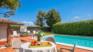 A poolside table with a fruit bowl, wine bottle, and glasses, surrounded by chairs, with a pool and greenery in the background. - Villa Oasi With Private Pool BBQ & Garden Near Sirmione (Sirmione)
