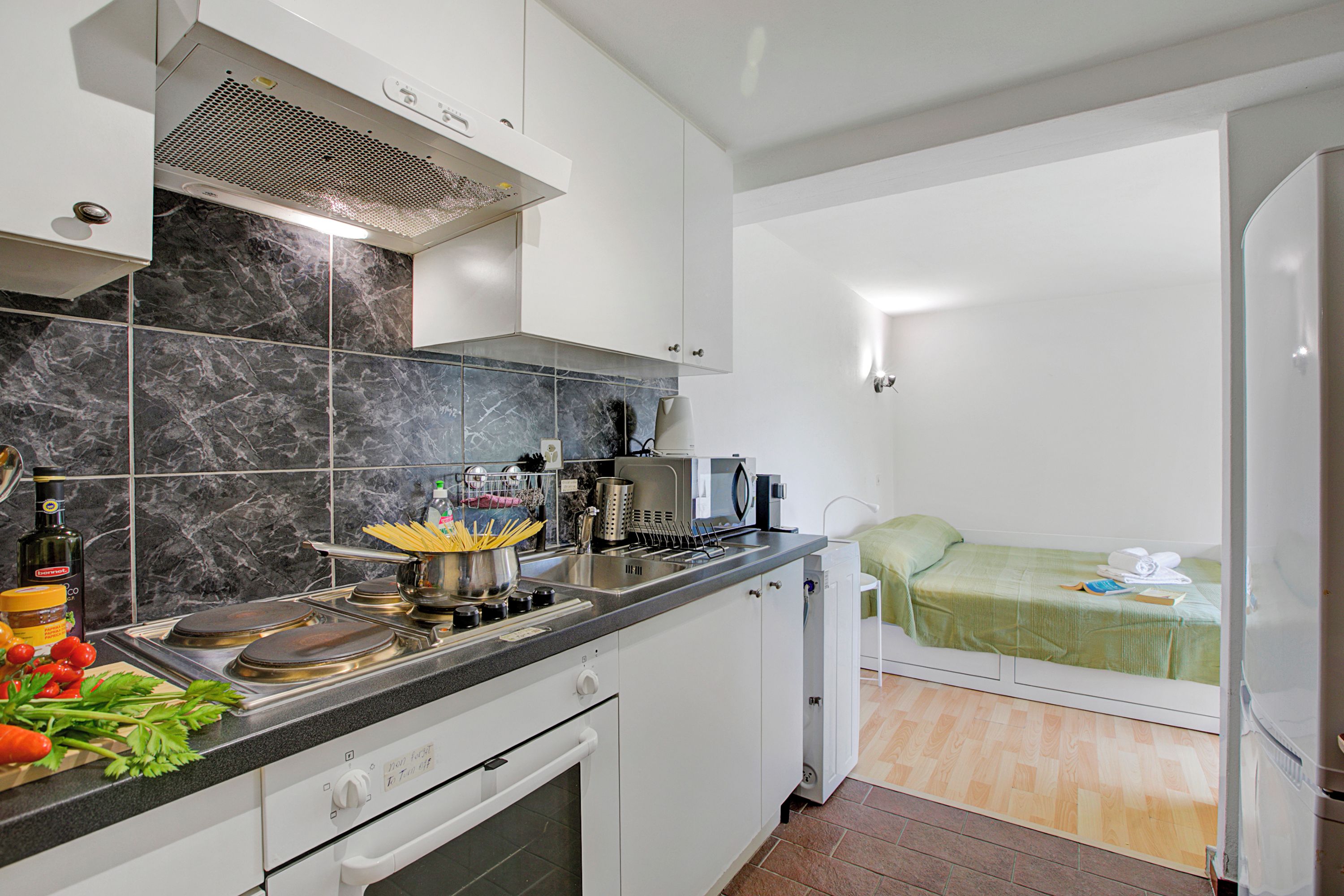 A compact kitchen with a stove, sink, and microwave. A bed is visible in the adjacent room.