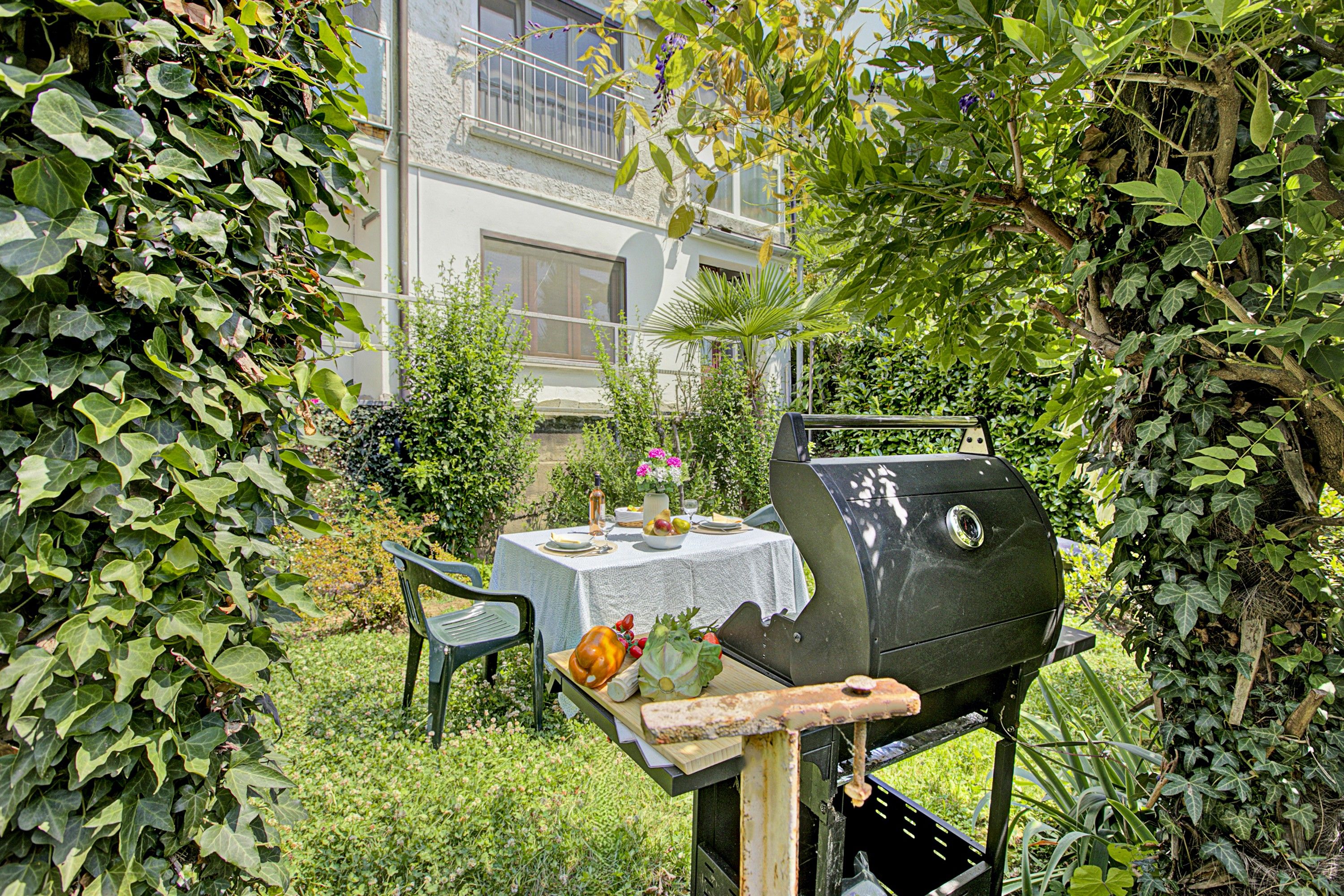 Patio trasero con una parrilla, una mesa preparada para una comida y una casa al fondo.