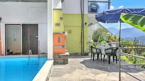 A swimming pool with a stone deck, a dining area with tables and chairs, and a view of mountains. - Studio W/panoramic View, Shared Pool, Garden & Terrace (Agno)