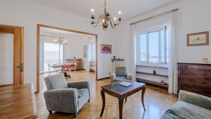 Un vaste salon doté d’un plancher en bois, d’un lustre, d’un coin repas avec une table et des chaises, et offrant une vue sur la mer grâce à de grandes fenêtres.