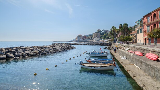 Une ville côtière où des bateaux sont amarrés le long d’une digue rocheuse, avec une promenade bordée de bâtiments et un ciel dégagé.