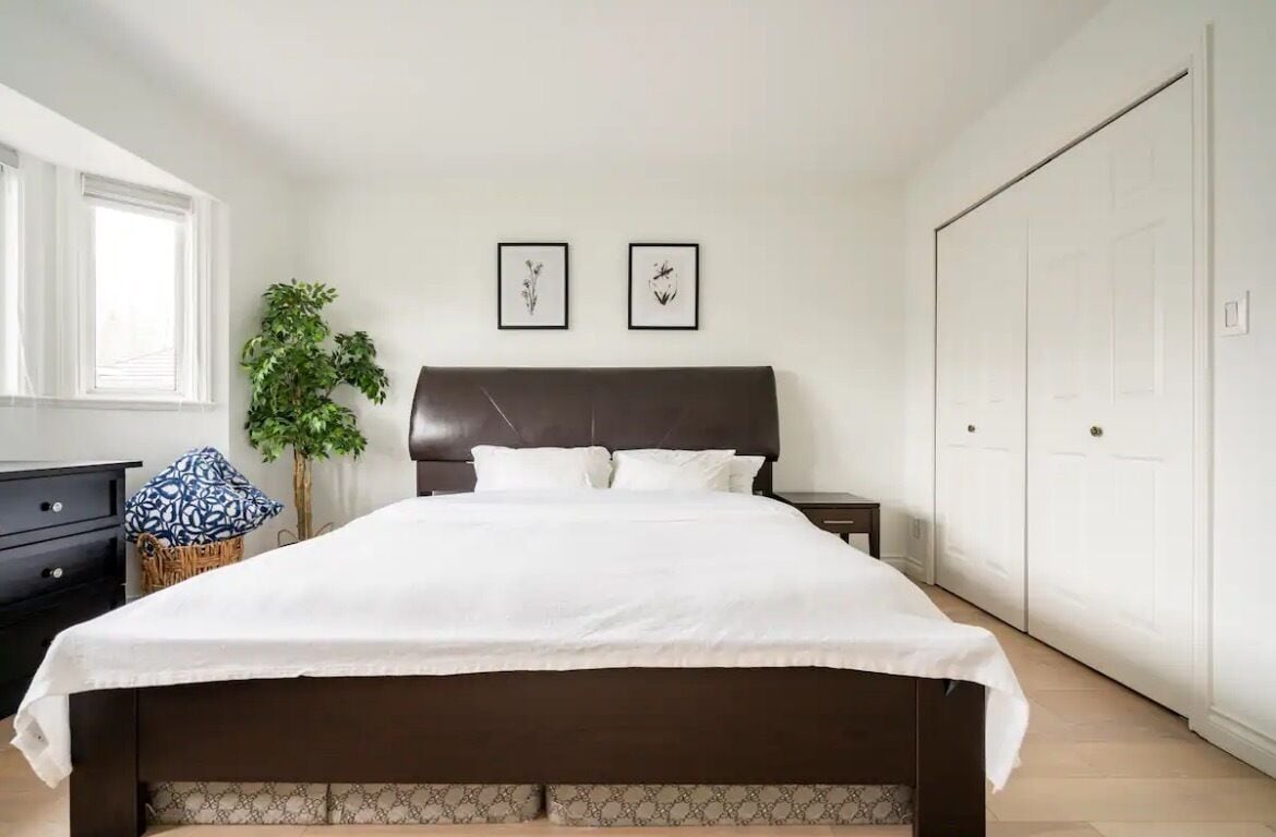 Un dormitorio con una cama grande, una mesita de noche, un aparador, una planta en maceta y dos cuadros en la pared.