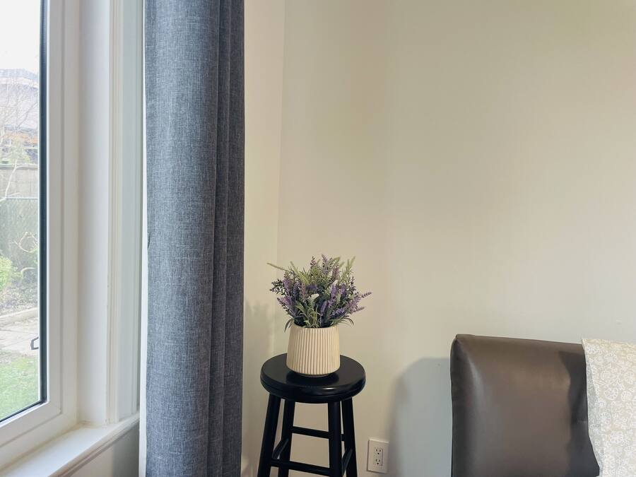 A room with a window, a grey curtain, a small stool with a plant, and a brown chair.